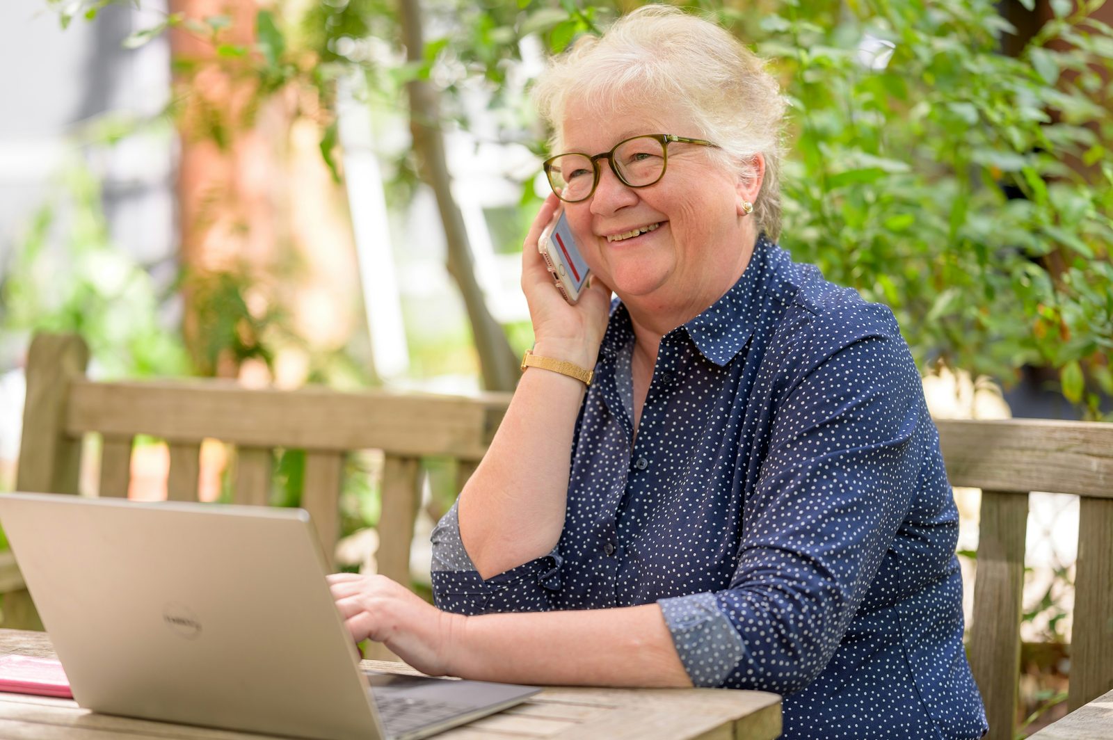 Smiling senior woman on a phone call in her garden with her laptop